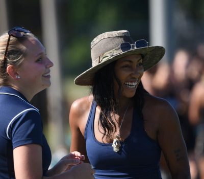 Two women smiling and talking outdoors on a sunny day. One is wearing a dark blue shirt with sunglasses on her head, and the other is wearing a wide-brimmed hat, sunglasses resting on the hat, a navy tank top, and a necklace. The background is blurred with other people visible.